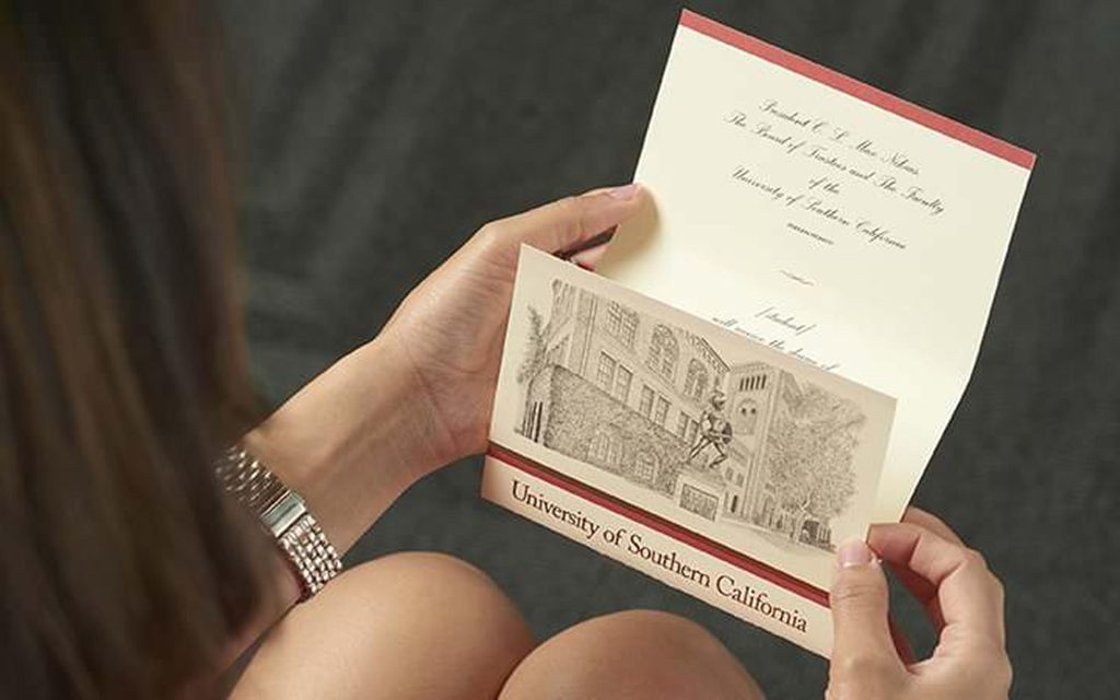 A person holding and reading an acceptance letter from the University of Southern California, featuring an illustration of a campus building on the front.
