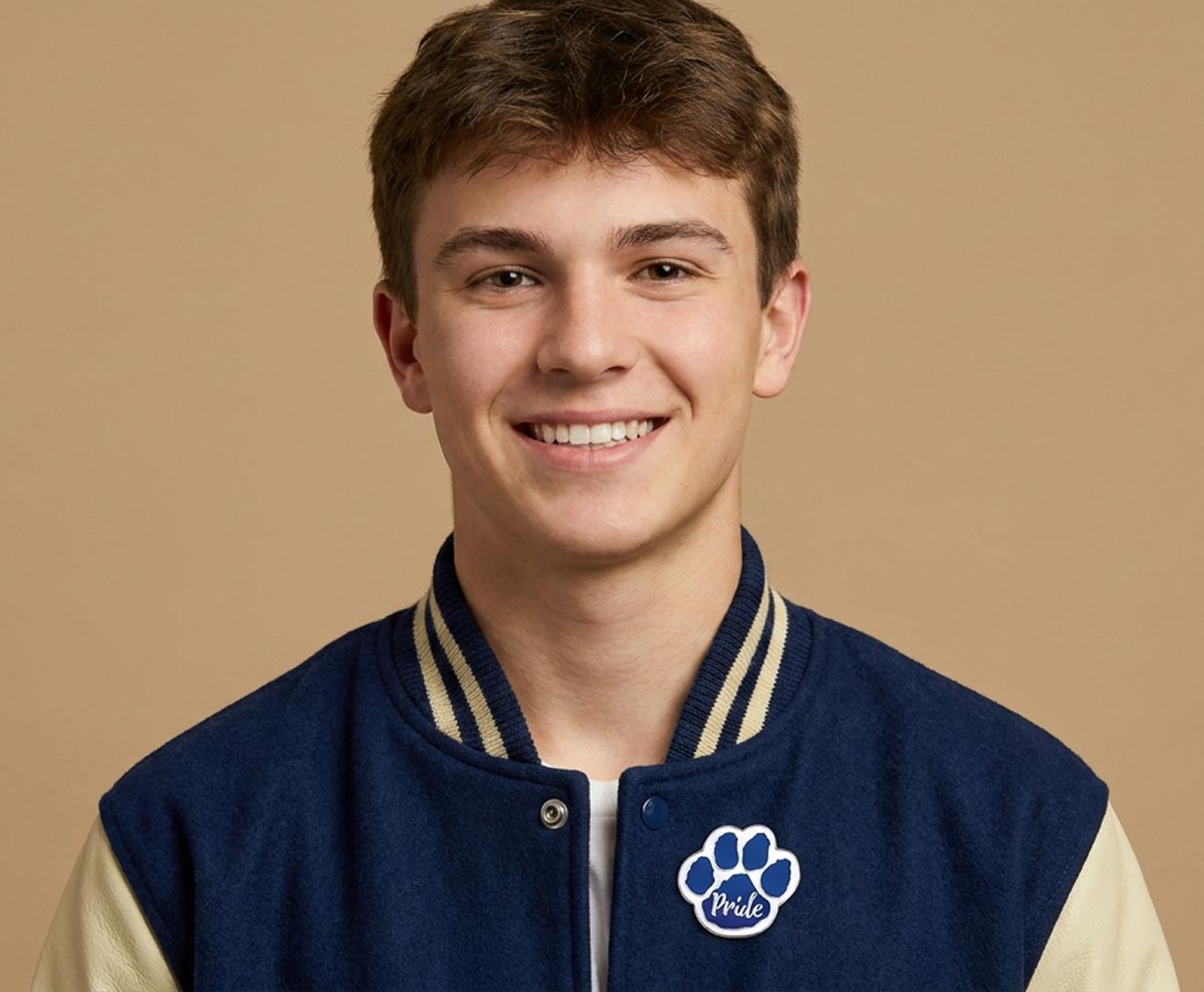 Smiling young person with short brown hair wearing a blue and cream varsity jacket with a paw print patch that says Pride, against a beige background.