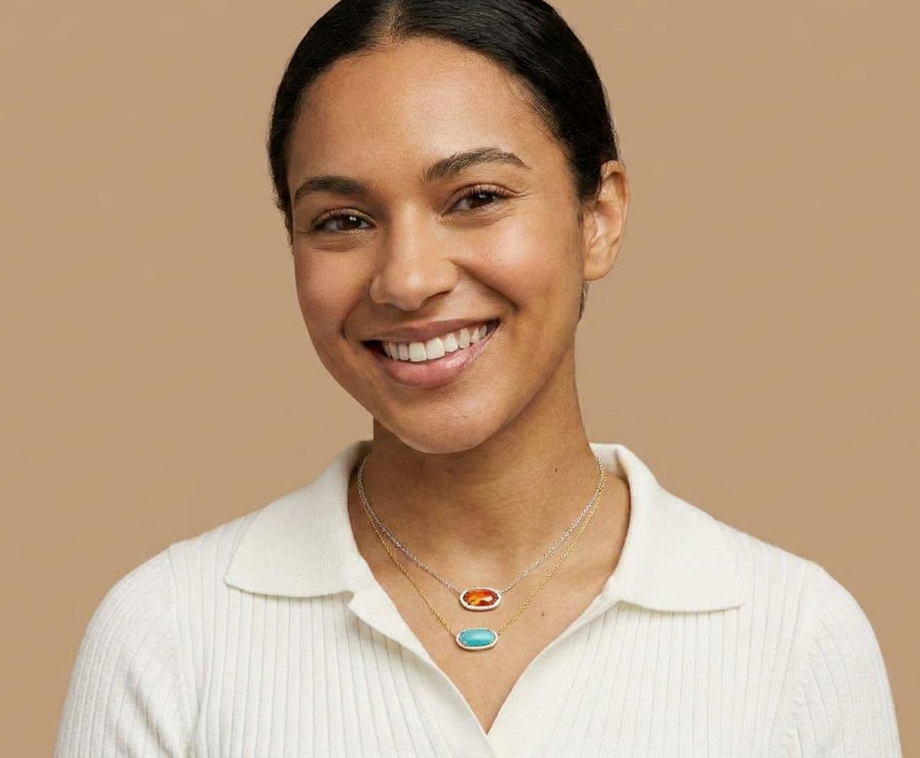 Smiling woman with dark hair tied back, wearing a white collared shirt and layered necklaces with orange and turquoise pendants, posed against a beige background.