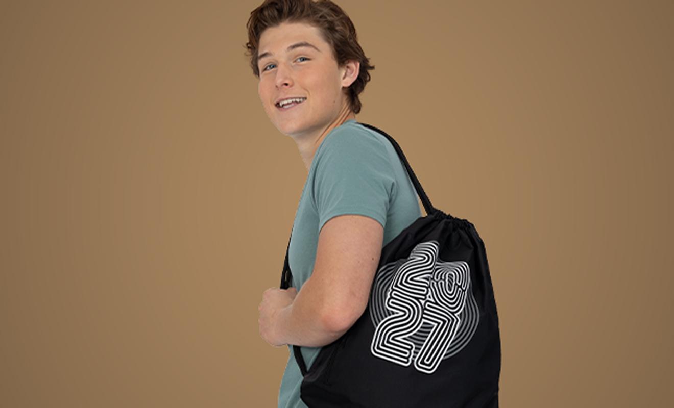 A young person with short brown hair, wearing a teal shirt, smiles while carrying a black drawstring bag with a bold white 2021 graphic on it, standing against a plain brown background.
