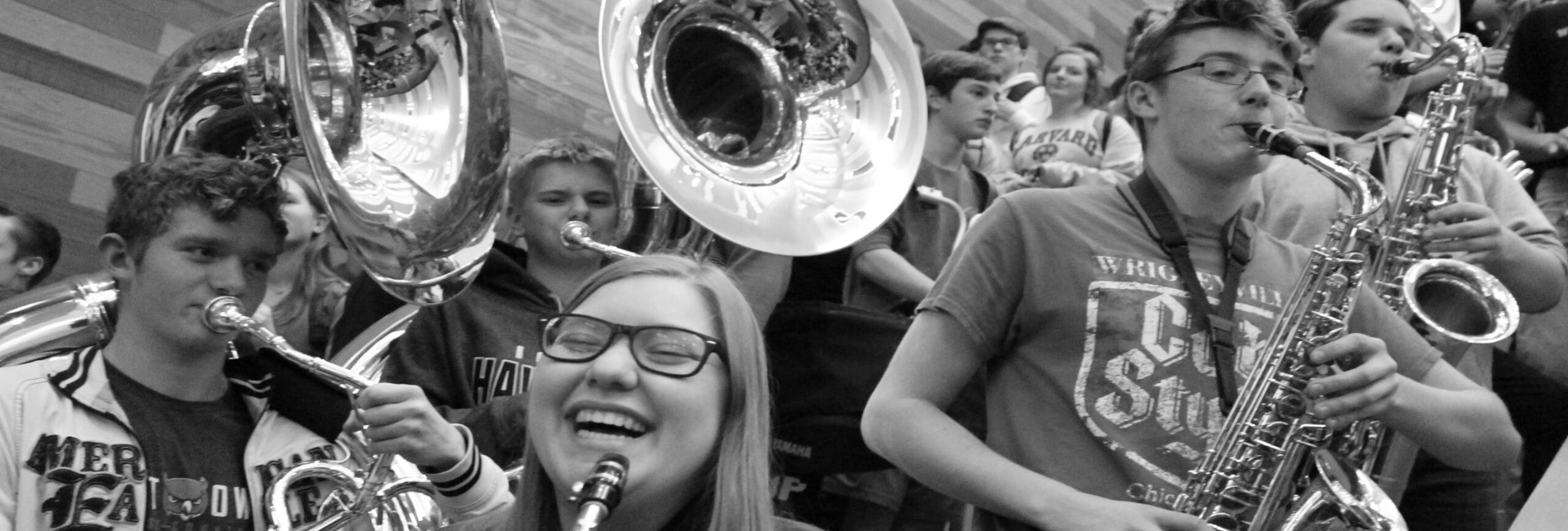 A group of students plays musical instruments, including saxophones and sousaphones, in a band. One student is smiling widely. The background shows more students and instruments. The image is in black and white.