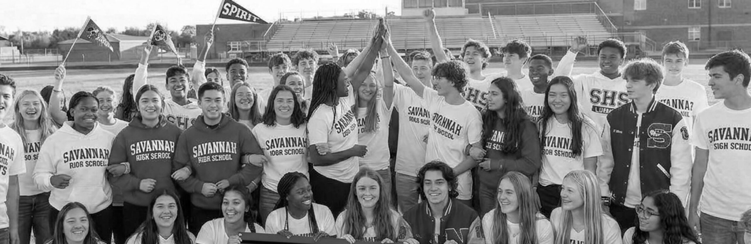 A large group of high school students in Savannah High School shirts gather outdoors, smiling and cheering together, some raising their arms and holding banners, on an athletic field.