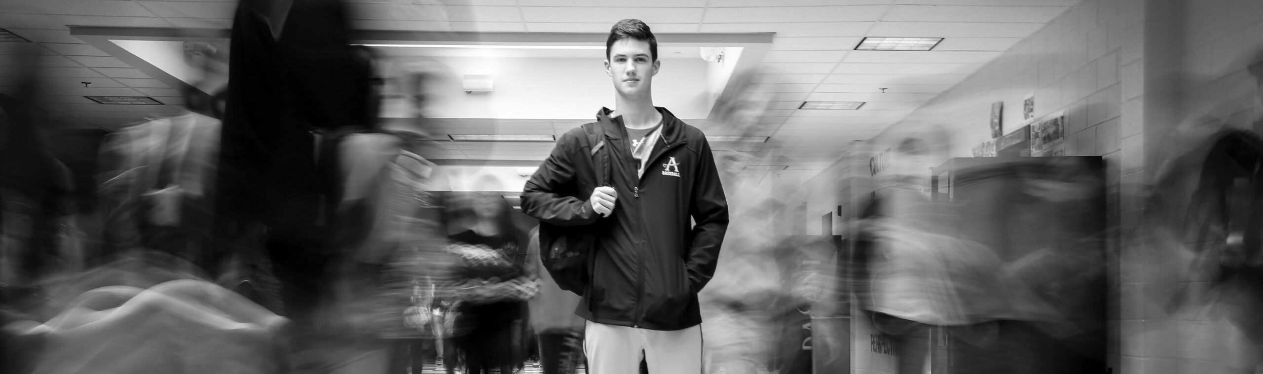 A young man stands still in a school hallway, looking at the camera, while blurred students walk past him on both sides, creating a sense of motion around his calm presence.