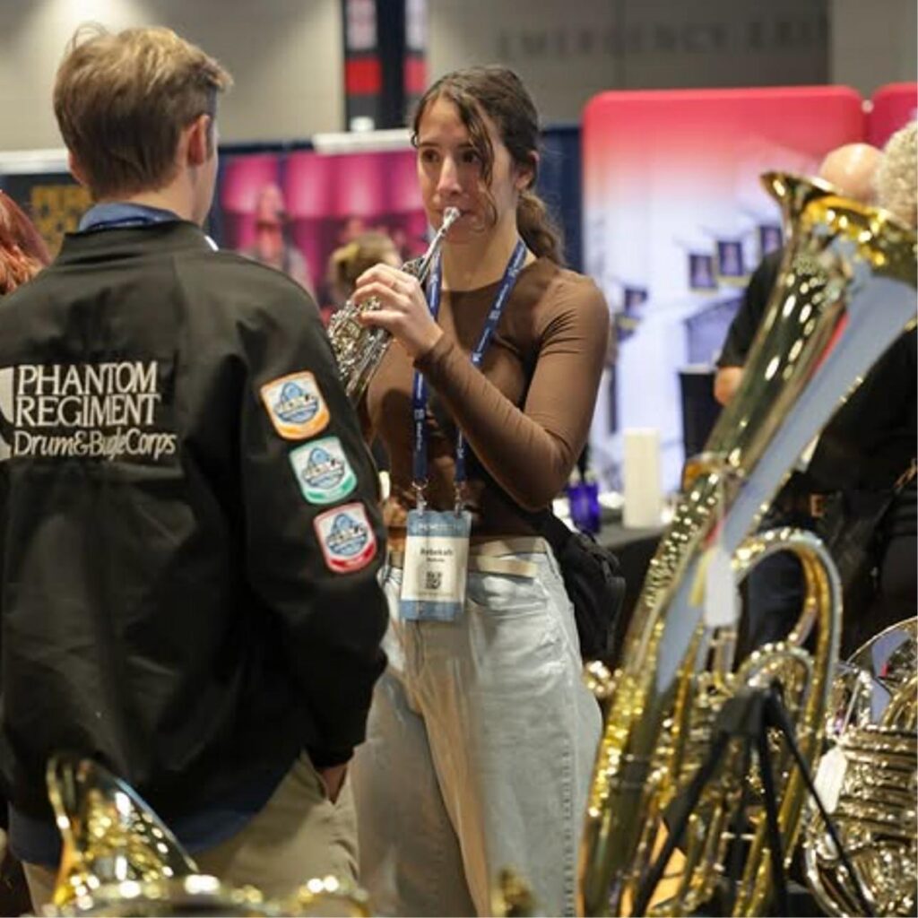 A woman plays a brass instrument at a music event, facing a man wearing a Phantom Regiment jacket. Shiny instruments are displayed around them, and people are visible in the background.