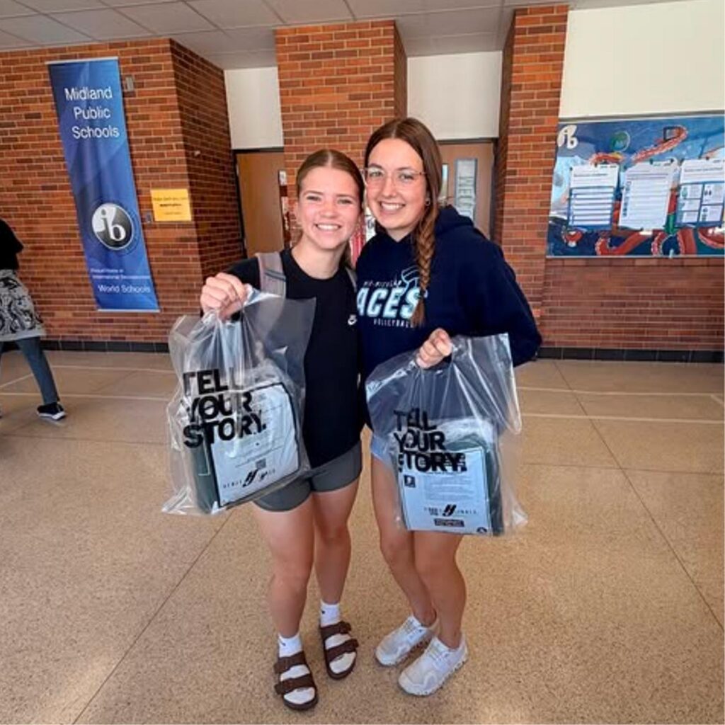 Two smiling students stand indoors holding plastic bags that say TELL YOUR STORY. The background features a brick wall, school banners, and a colorful mural.
