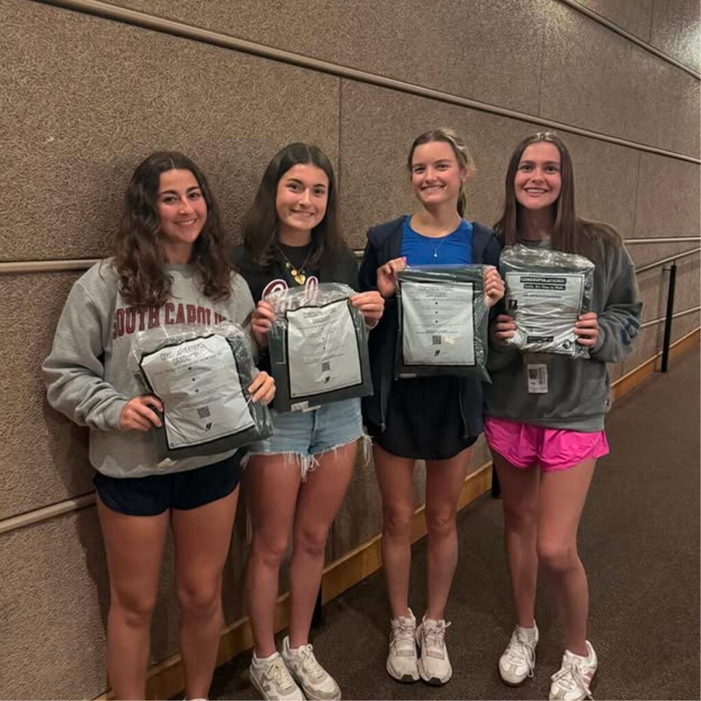 Four young women stand indoors by a textured wall, smiling and holding clear plastic bags containing folded clothing items. They are dressed casually in shorts, T-shirts, and sneakers.