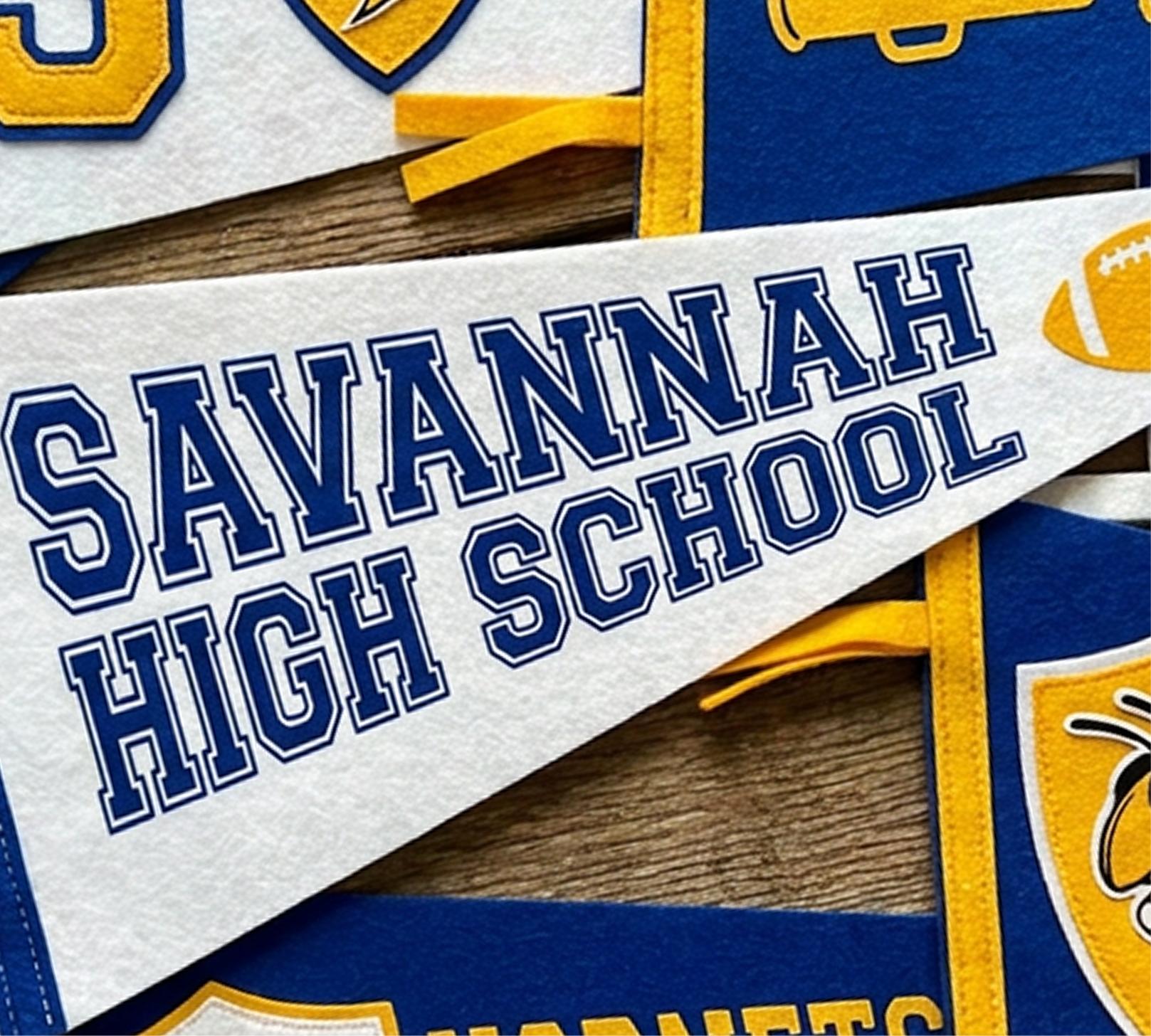 A white felt pennant reads “SAVANNAH HIGH SCHOOL” in blue letters, with a yellow football graphic. Blue and yellow banners with athletic motifs are partially visible underneath.