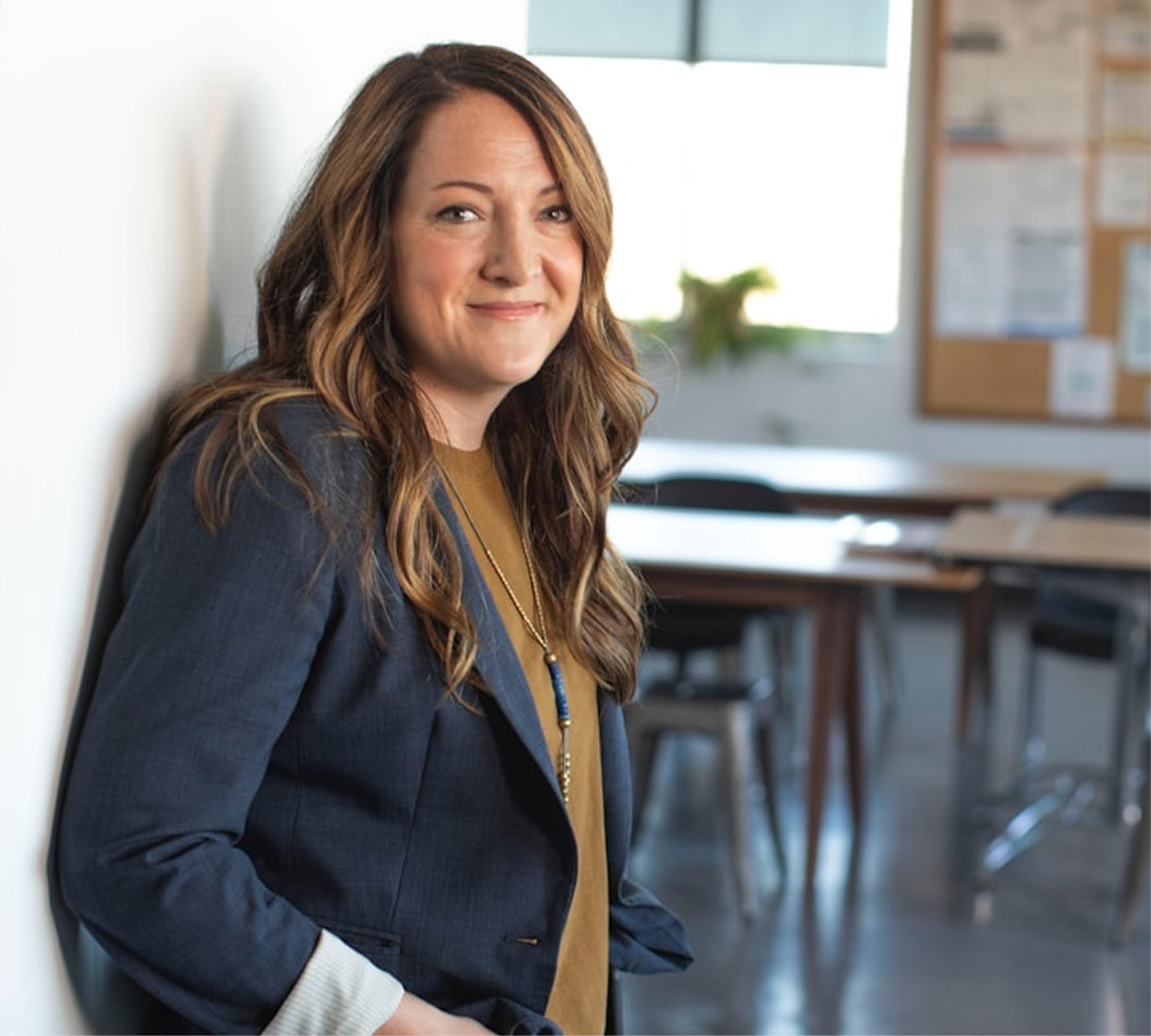 A woman with wavy brown hair, wearing a blue blazer and tan shirt, smiles while leaning against a white wall in a classroom with tables, chairs, and a bulletin board in the background.