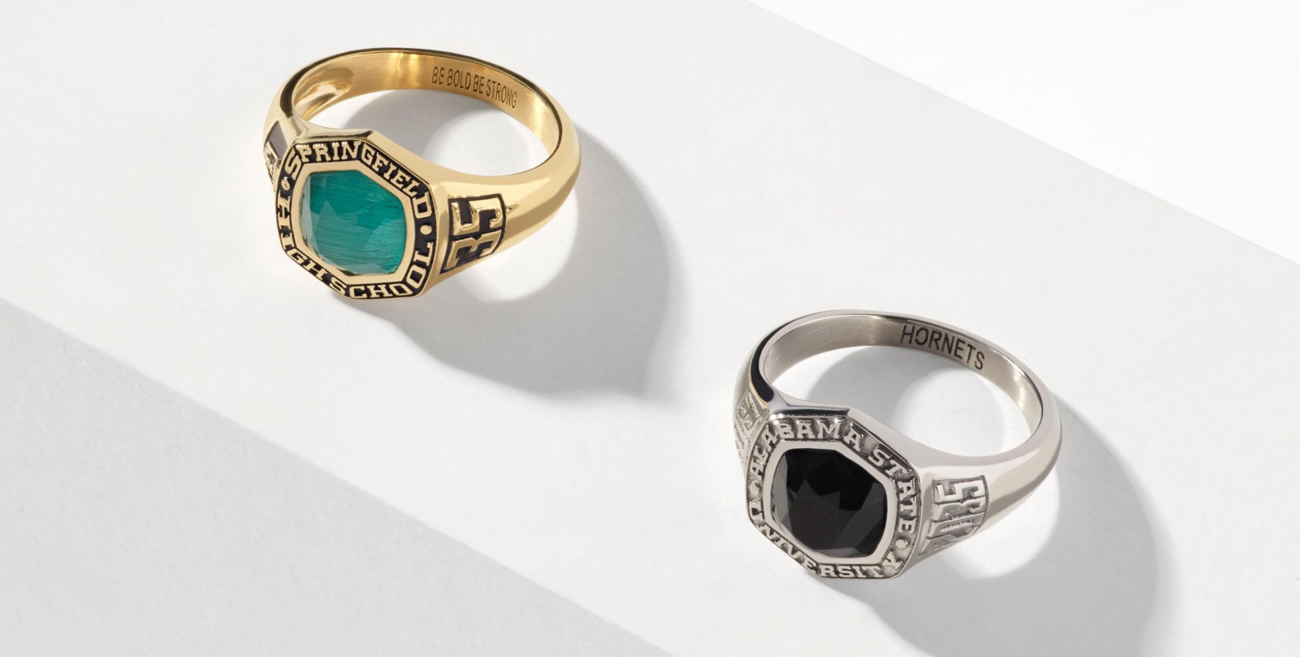 Gold and silver class rings rest on a white surface. The gold ring features a green gemstone, while the silver ring has a black gemstone. Both rings have engraved details on their bands and around the stones.