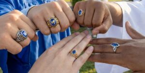 Five hands, each wearing a gold or silver ring with a blue stone, are joined together in a circle; some hands wear blue graduation gowns.