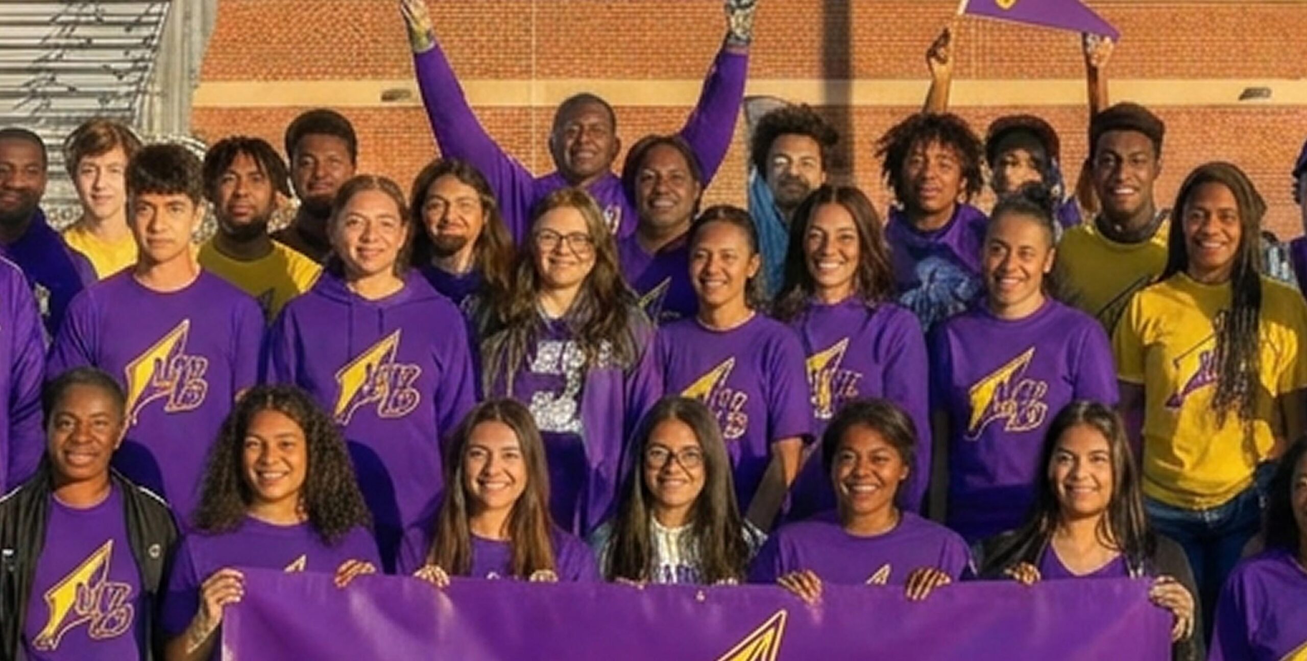 A group of students wearing purple and yellow shirts with a lightning bolt logo pose together outside, smiling and holding a matching purple banner.