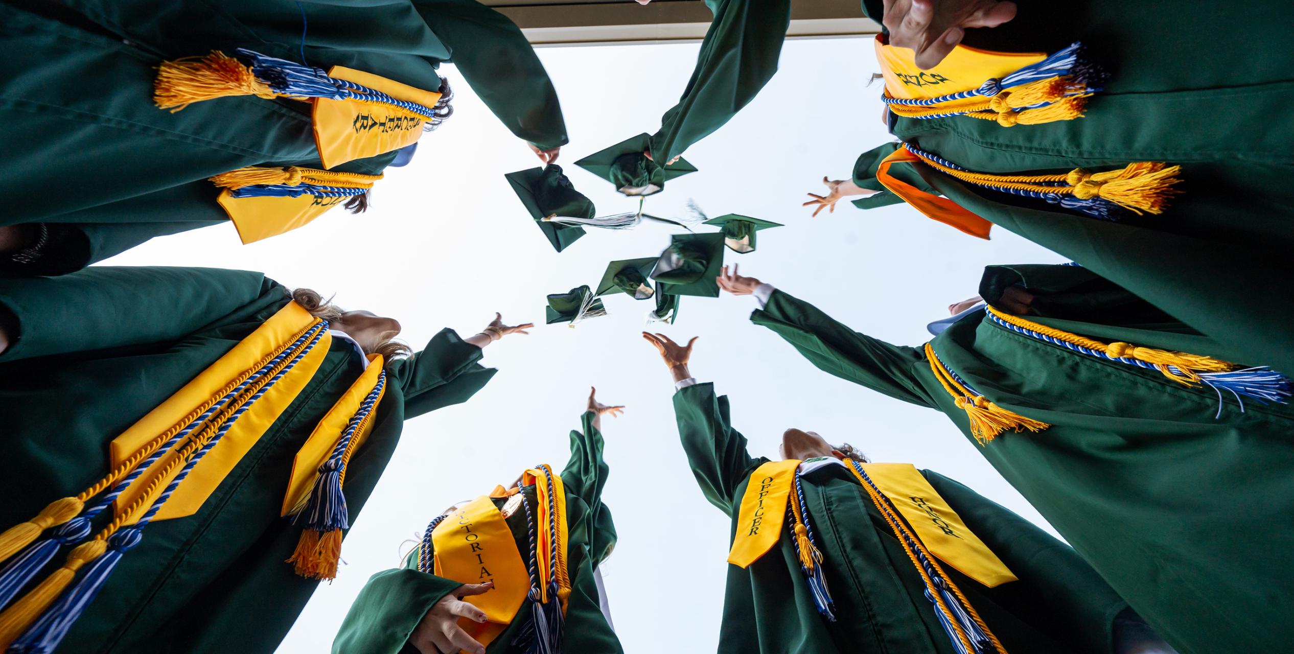 Graduates in green robes and yellow stoles stand in a circle, tossing their caps into the air, celebrating against a bright sky, as seen from a low-angle perspective.